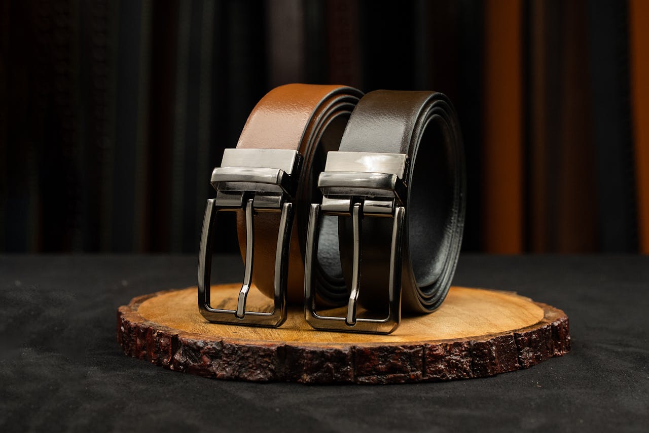 Two stylish leather belts with metal buckles displayed on a wooden platform against a striped backdrop.